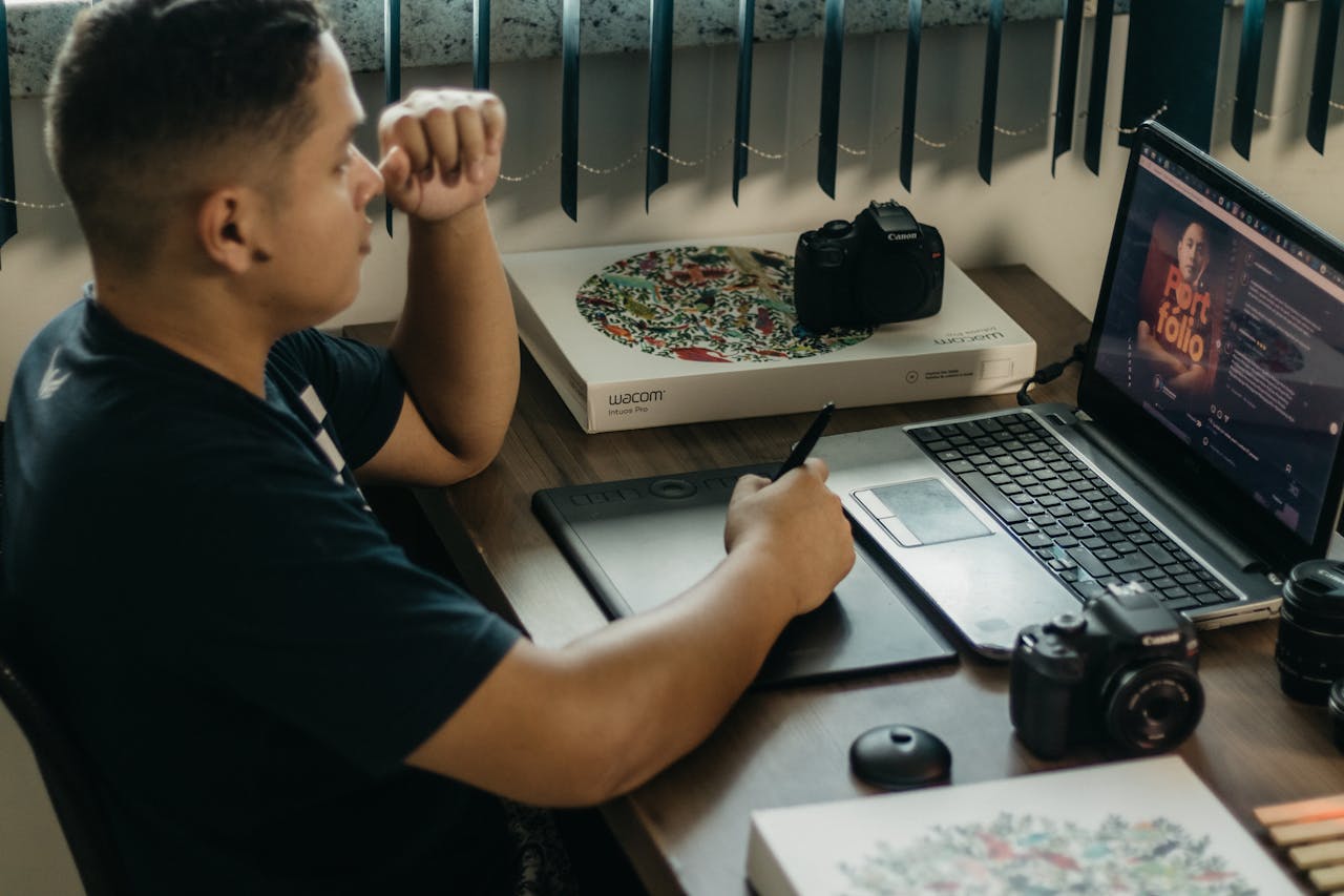 A photographer working at a creative setup with a laptop and Wacom tablet, focused on editing photos indoors.